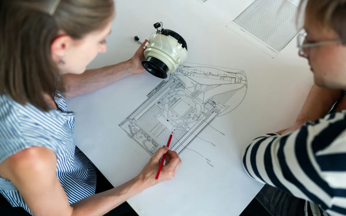 Zwei Ingenieurinnen prüfen eine technische Zeichnung und ein Bauteil am Tisch